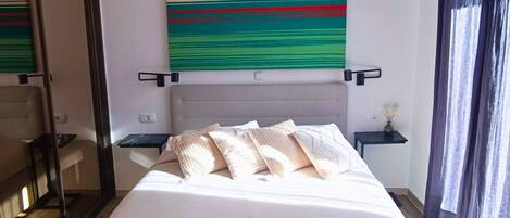 Habitación de hotel moderna con una cama grande, un espejo, una mesita de noche con lámpara y un cuadro abstracto y colorido en la pared.