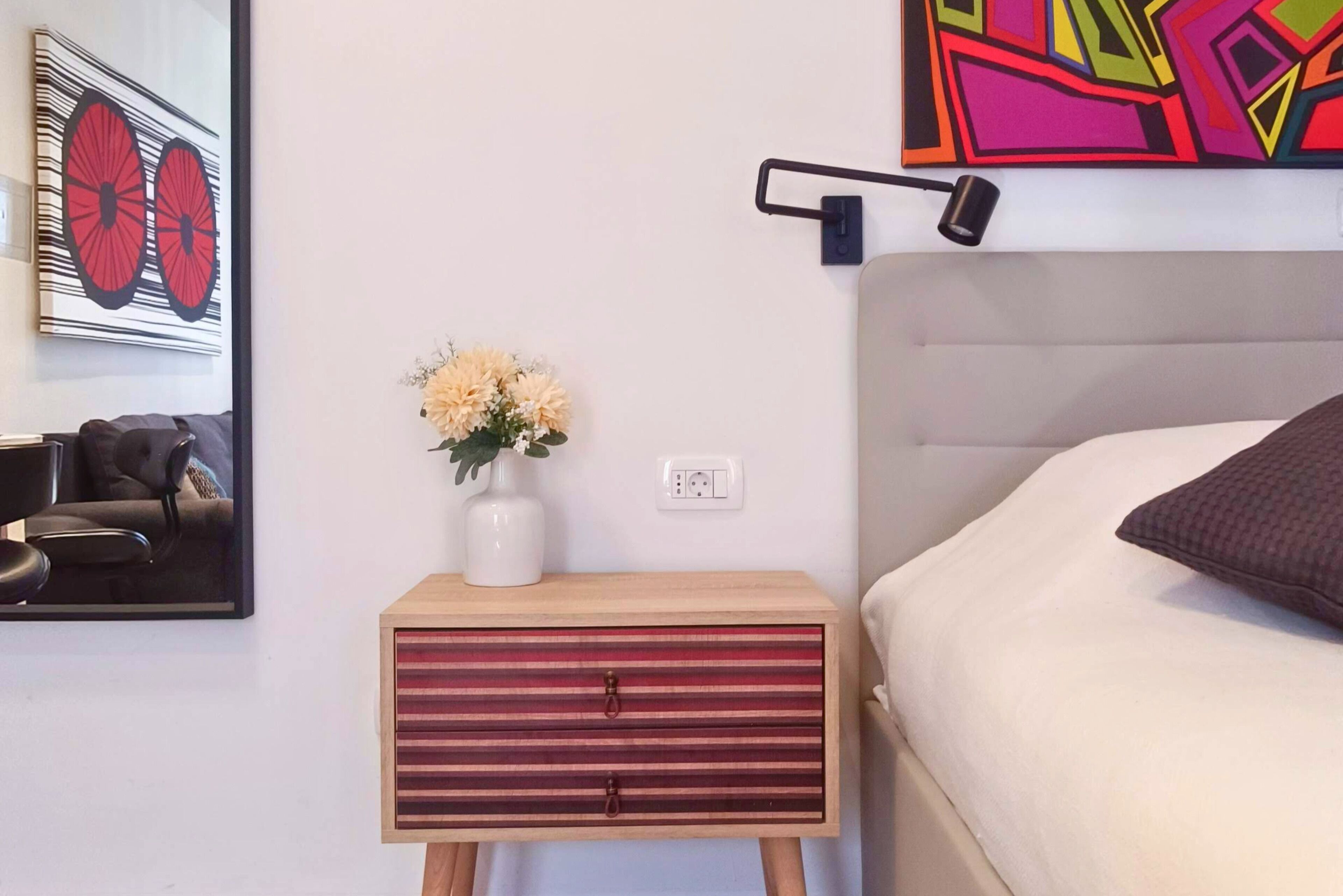 A hotel room with a bed, a nightstand with a vase of flowers, a chair, and a wall art piece.
