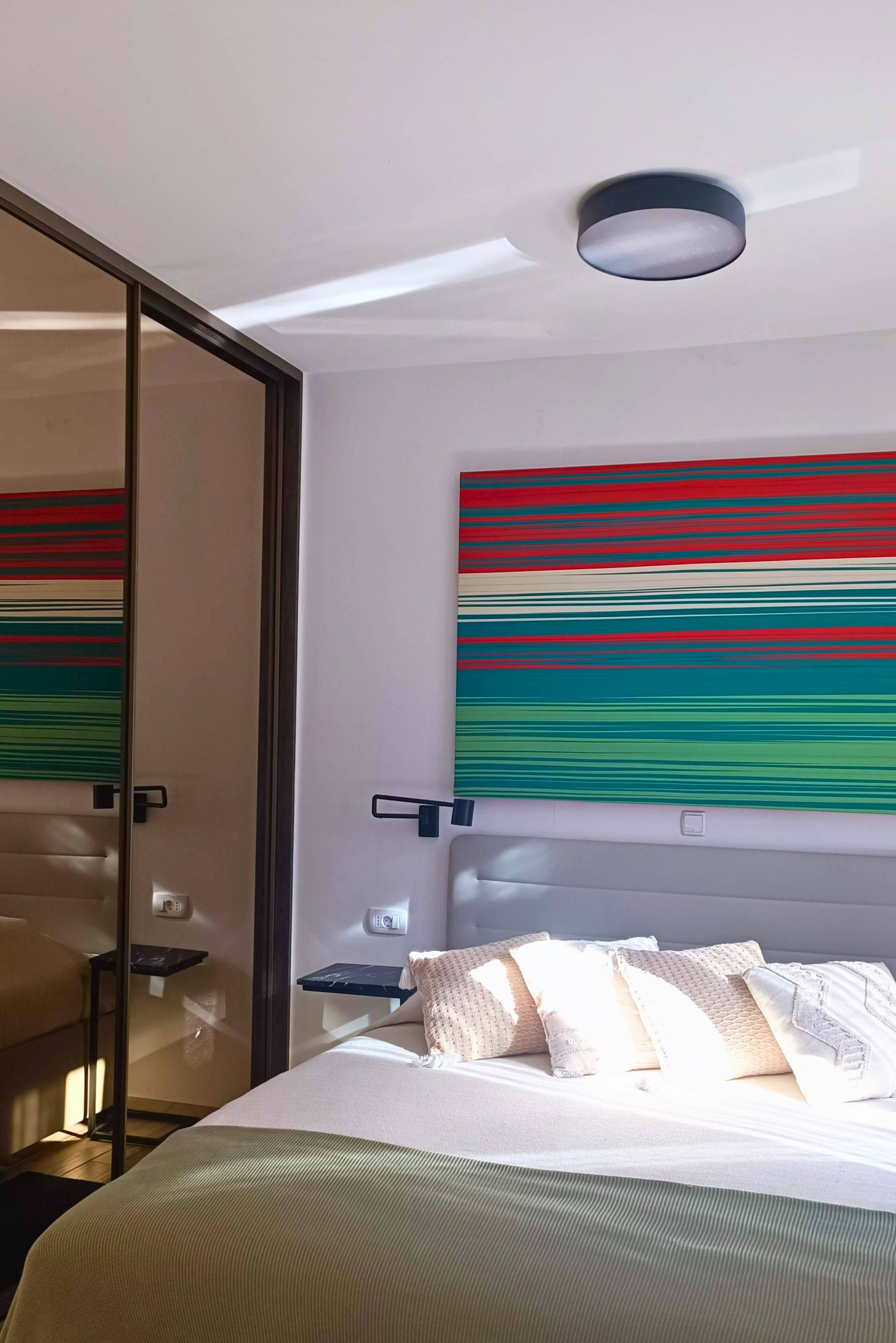 A modern hotel room with a bed, a bedside table, a wall-mounted artwork, and a ceiling light.