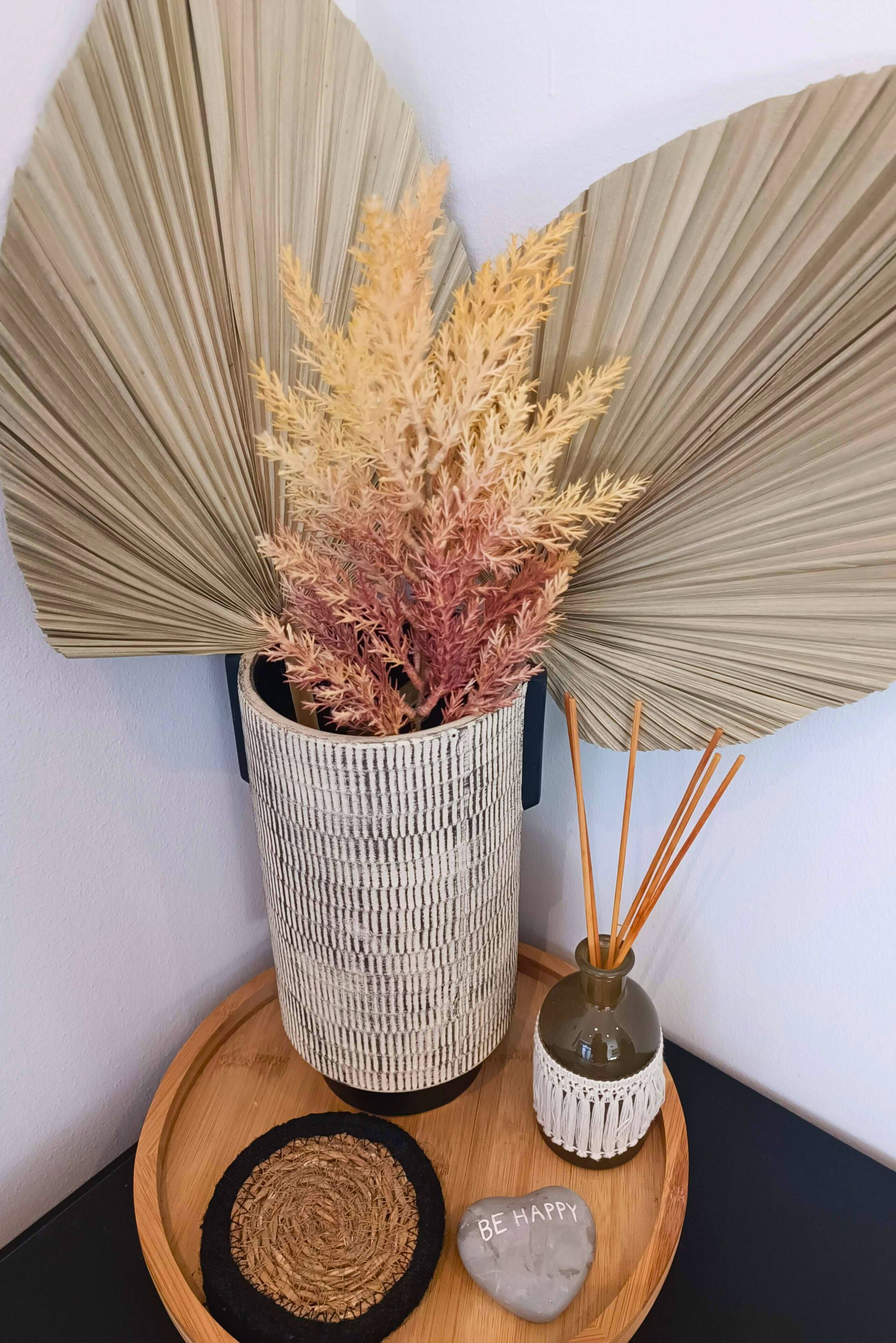 A decorative arrangement with a woven pot containing dried plants, a small vase with reed sticks, and a round wooden tray with a woven mat and a stone with the inscription "BE HAPPY".