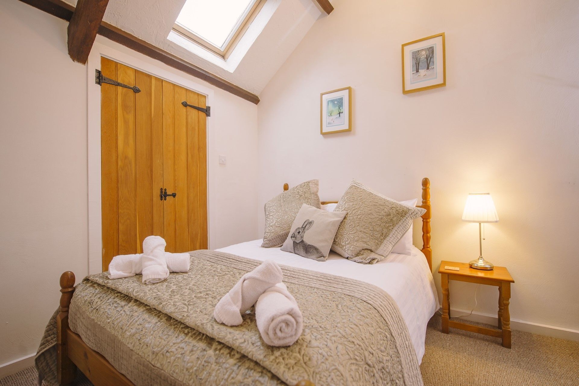 A bedroom with a wooden door, a bed with rolled towels, a bedside lamp, and two framed pictures on the wall.