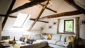 Ein gemütliches Loft-Zimmer mit freiliegenden Holzbalken, einem großen grauen Ecksofa, einem Esstisch mit Stühlen und einem Kronleuchter.