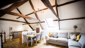 Ein gemütlicher Loft-Raum mit freiliegenden Holzbalken, einer Essecke mit Holztisch und -stühlen, einer Couch mit Kissen und einer kleinen Küchenzeile im Hintergrund.