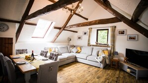 Ein gemütlicher Loftsraum mit freiliegenden Holzbalken, einem großen Fenster, einem Ecksofa, einem Esstisch für zwei Personen und einem Fernseher auf einem Holzständer.