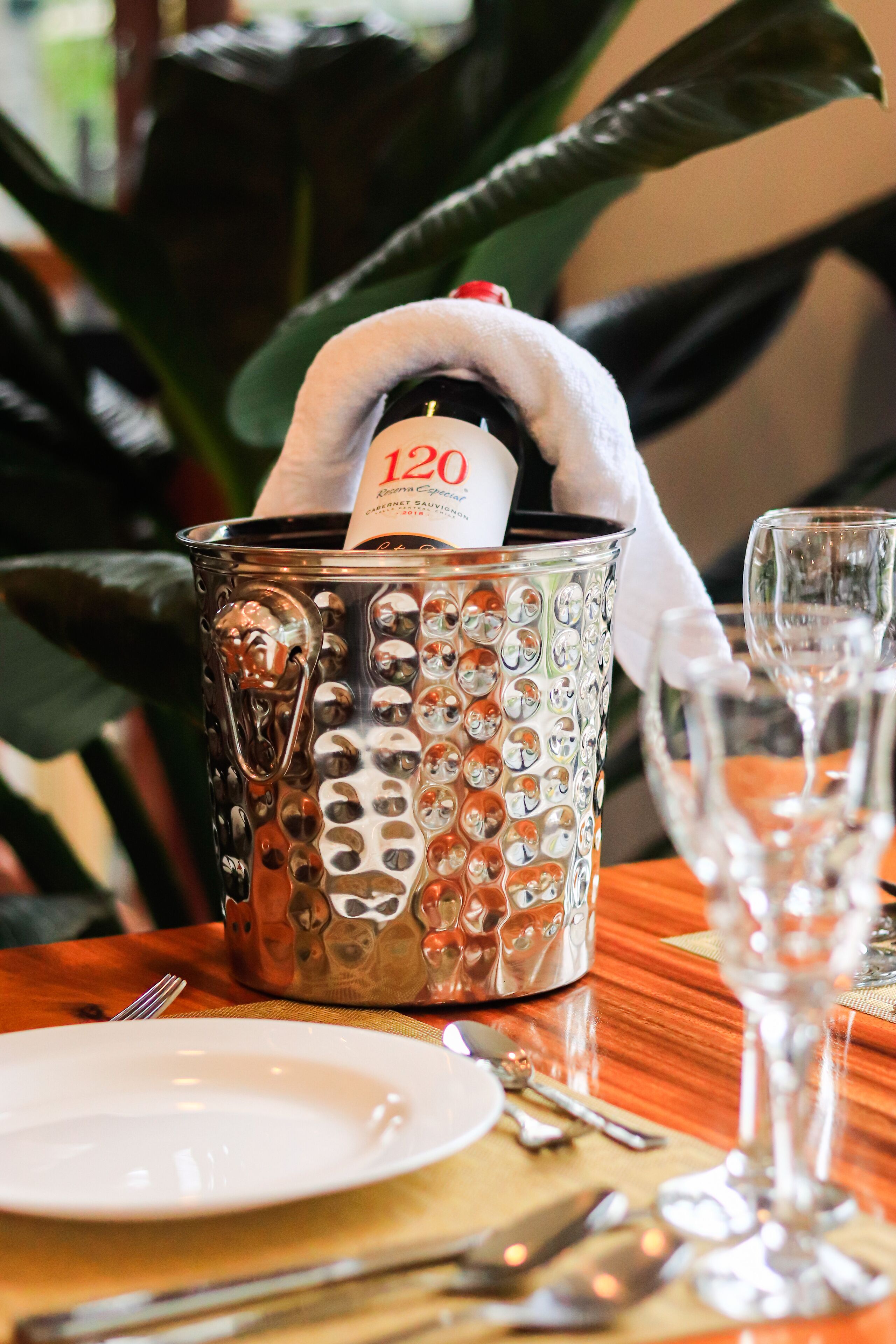 A table setting with a champagne bottle in a decorative metal container, a white plate, and a wine glass.
