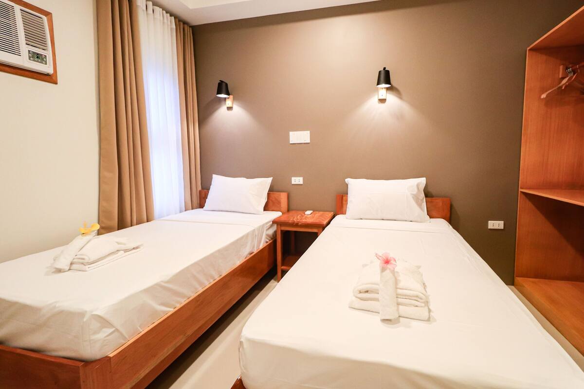 A hotel room with two single beds, a wooden wardrobe, a small table, and a wall-mounted heater.