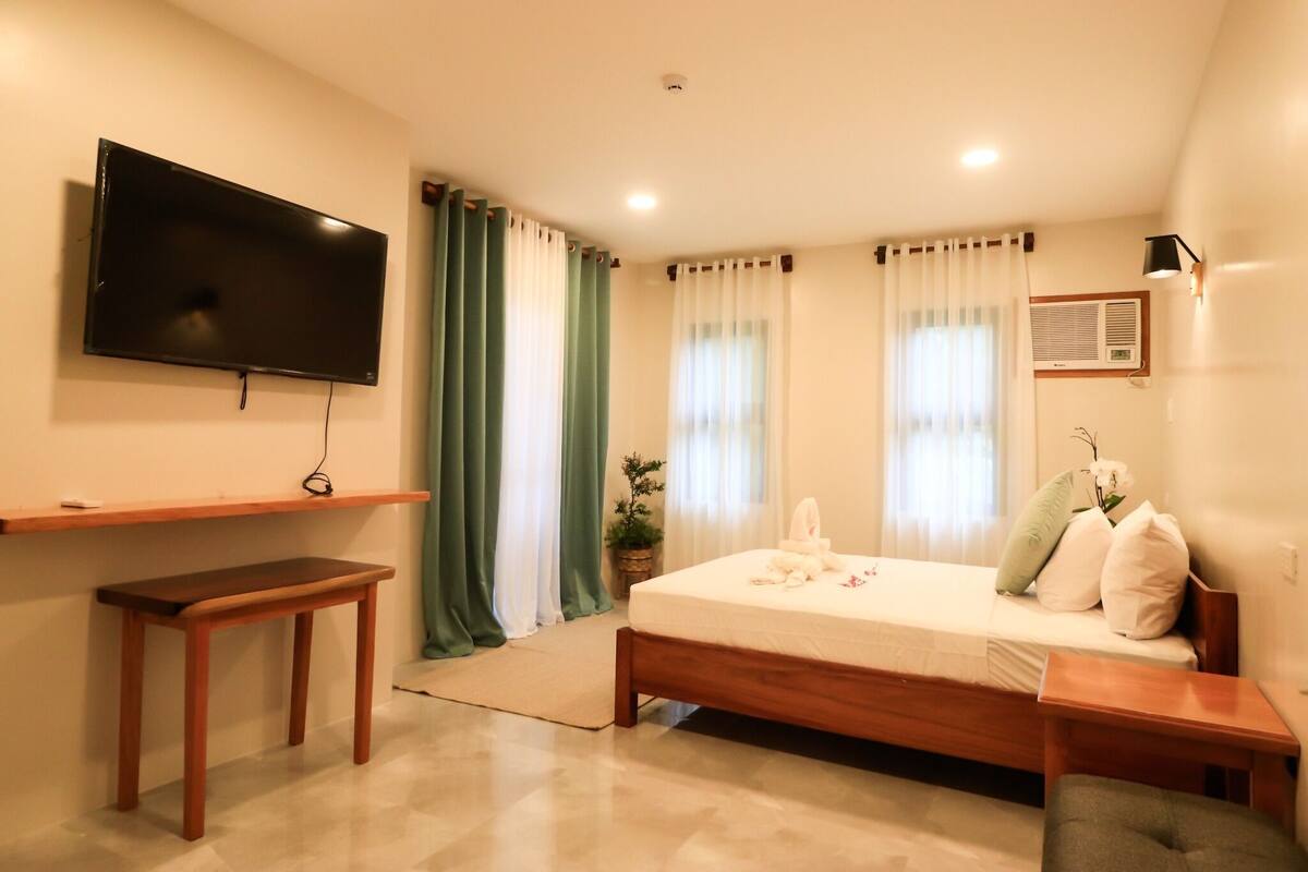 A bedroom with a bed, a TV mounted on the wall, a bedside table, a bench, and a potted plant.