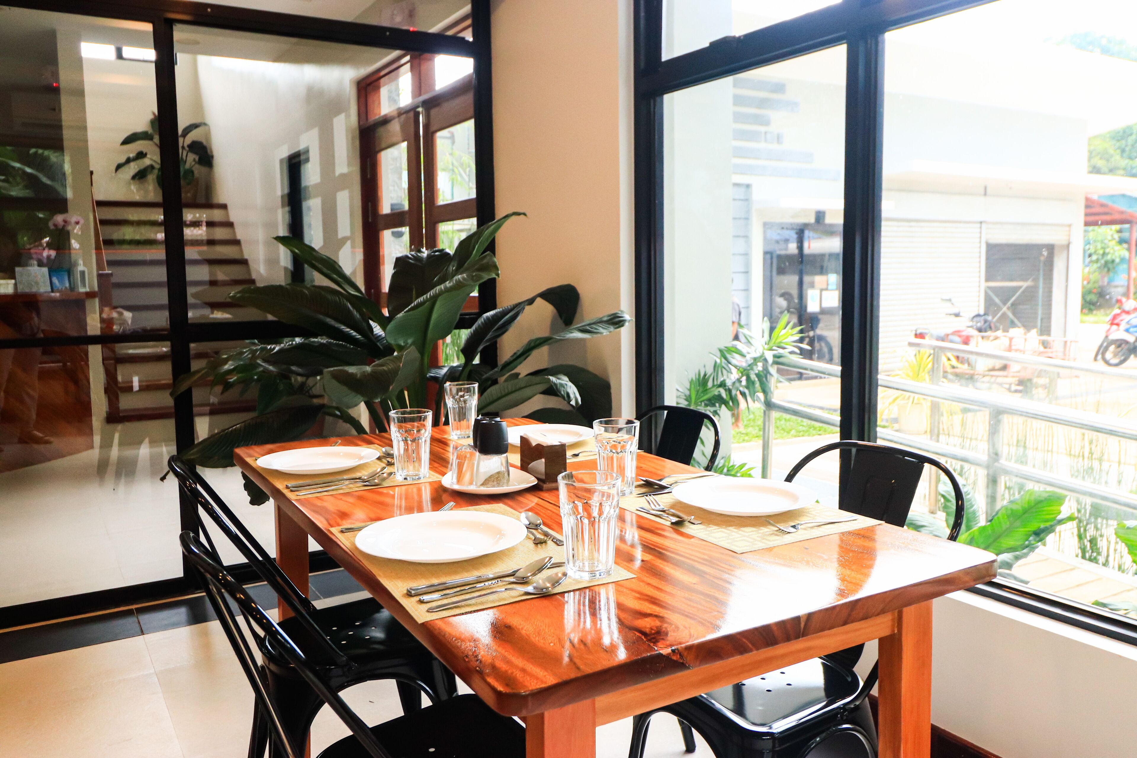A dining table set with plates, cutlery, and glasses, surrounded by chairs, with a view of a street and greenery outside.