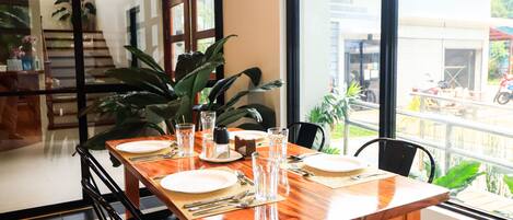A dining table set with plates, cutlery, and glasses, surrounded by chairs, with a view of a street and greenery outside.