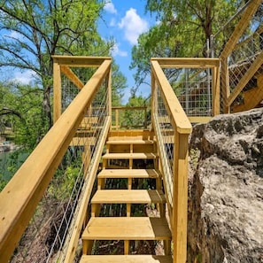 Una escalera de madera que sube a una plataforma, rodeada de árboles y una roca.