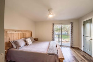 Un dormitorio con cabecera de madera, cama con colcha color beige, un banco al pie de la cama, ventilador de techo y una puerta corrediza de vidrio que da a un espacio exterior.