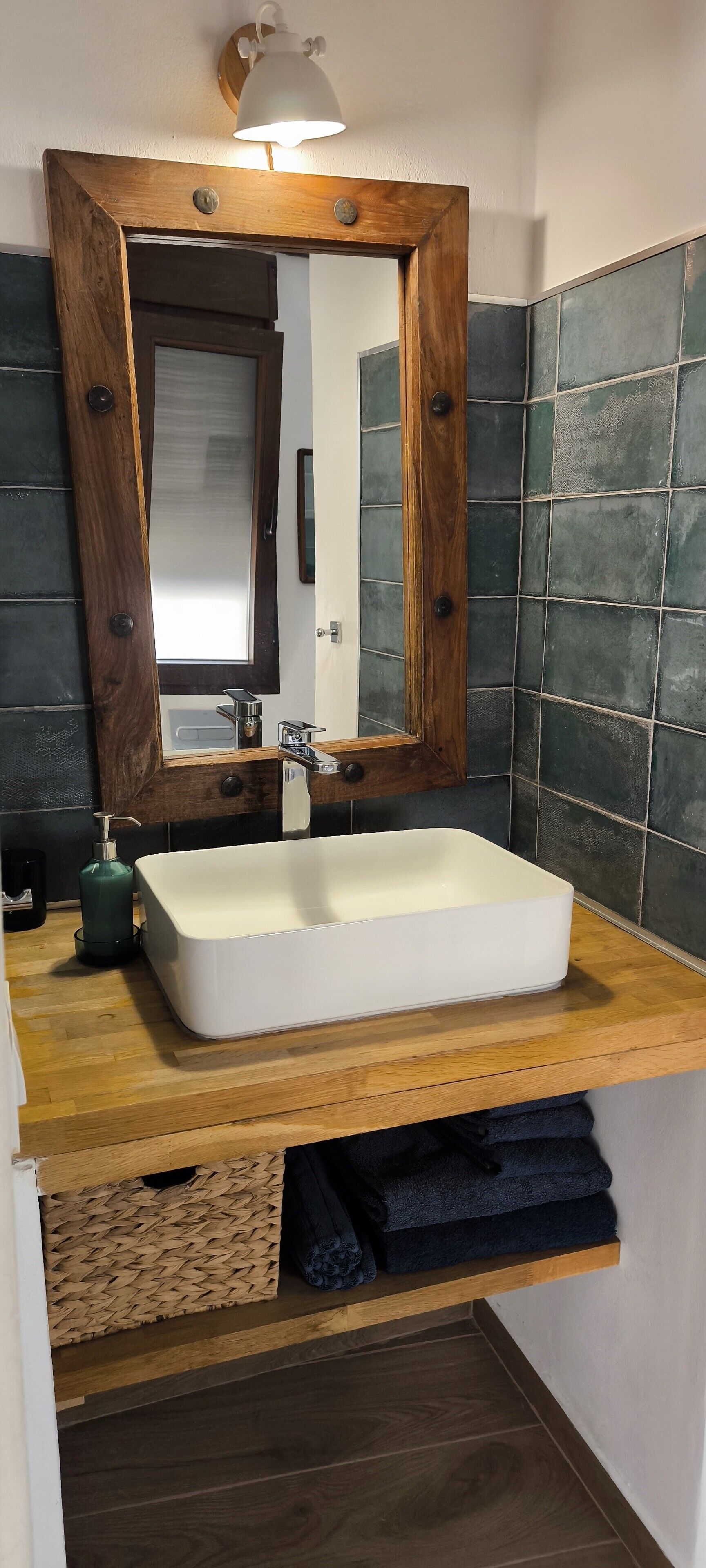 Une salle de bain avec un meuble-lavabo en bois, un lavabo blanc, un miroir avec un cadre en bois et des murs recouverts de carreaux foncés.