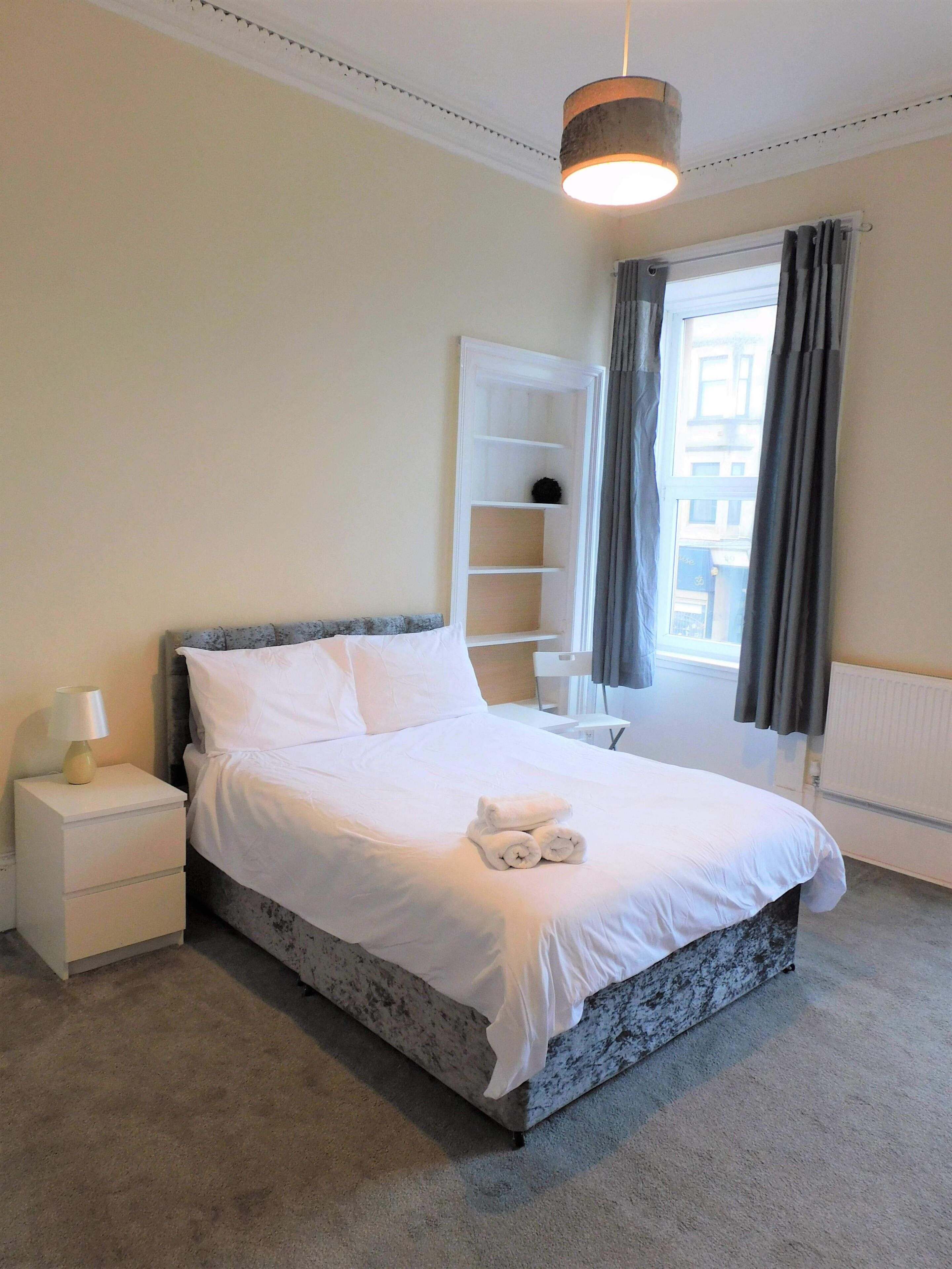 A neatly made bed with white linens, a bedside lamp, and a small shelf unit.
