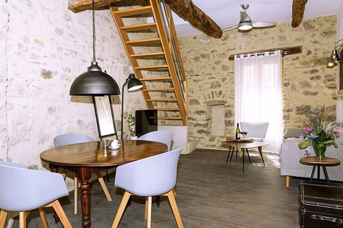 A rustic room with a wooden dining table, white chairs, a ladder, a fireplace, and a small round table with a vase of flowers.