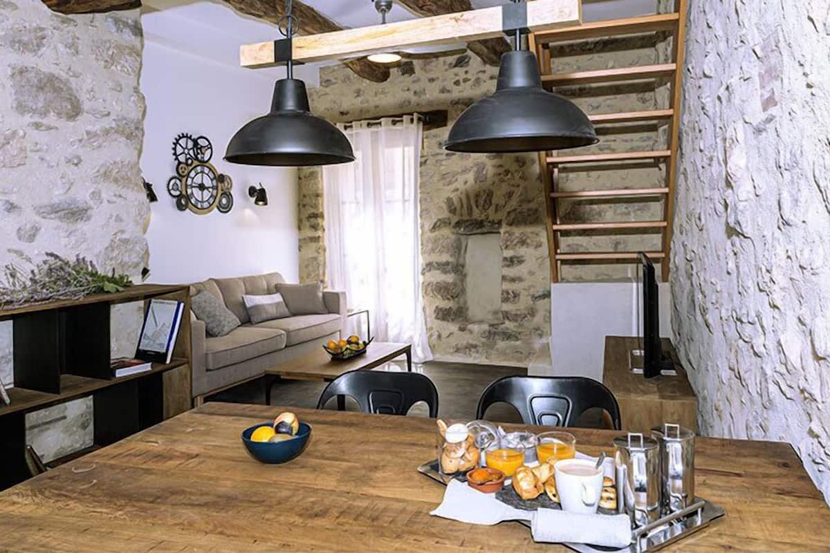 A rustic interior with a wooden dining table set with breakfast items, a sofa, and a staircase with a wooden railing.