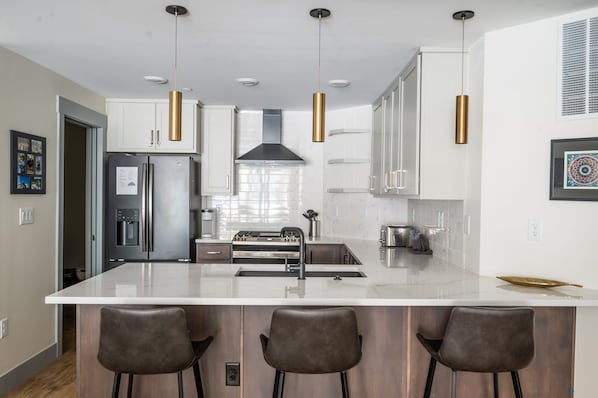 Une cuisine moderne dotée d’un îlot central, d’appareils électroménagers en acier inoxydable, de luminaires suspendus et d’une grande fenêtre.