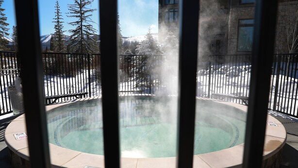 A hot tub with a stone surround, surrounded by a metal fence, with snow-covered trees and mountains in the background.