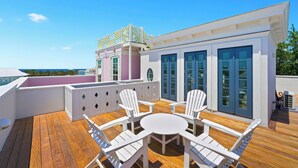 Um deck de madeira com cadeiras e uma mesa brancas, um edifício com portas azuis e vista para o oceano.