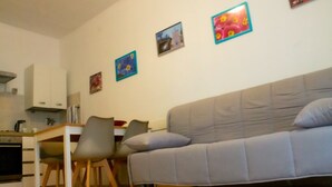 Ein Wohnzimmer mit grauem Sofa, zwei Sesseln, einem kleinen Tisch und einer Küchenzeile mit Schränken und Herd.