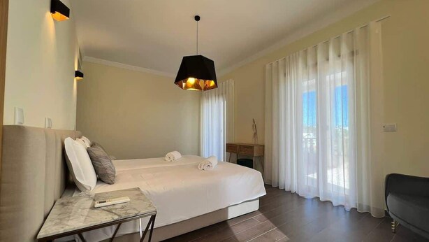 Una camera d'albergo con un letto, un tavolino con il telecomando, una poltrona e un'ampia finestra con tende.