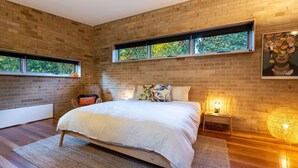 Ein modernes Schlafzimmer mit einem großen Bett, einem Sessel, einem Nachttisch und einem Fenster mit Blick auf Bäume.