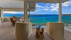 Une pièce dotée d’une grande fenêtre donnant sur une mer turquoise et des montagnes lointaines. On y trouve deux fauteuils beiges et une table en verre surmontée d’une sculpture de dauphin.
