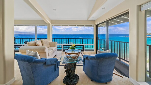 Un balcon avec vue sur l’océan, un canapé blanc, deux fauteuils bleus et une table basse en verre agrémentée d’un objet décoratif.