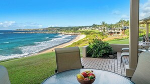 Une vue sur une plage avec des vagues de l’océan, une zone herbeuse et une table sur laquelle se trouve un saladier rempli de fruits.