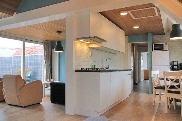 Een moderne keuken met een centraal eiland, witte kasten en een houten plafond. Er is een eethoek met stoelen en een tafel, en een zithoek met een bank en salontafel.