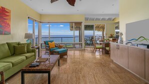 A spacious living area with a green sofa, a wooden coffee table, and a view of the ocean through large sliding glass doors.