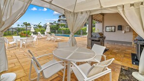 Eine Außenpatio-Terrasse mit weißen Metallstühlen und -tischen, einem Grill und einem Pool im Hintergrund.