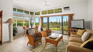 A spacious living room with a large sliding glass door leading to a balcony, wicker furniture, a flat-screen TV, and a ceiling fan.