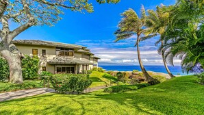 A two-story house with a large balcony, surrounded by lush greenery and palm trees, overlooking a clear blue ocean.