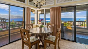A dining area with a round table and chairs, large windows with curtains, and a view of the ocean.
