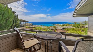 Un balcon avec une table et des chaises, offrant une vue sur la côte, avec des bâtiments aux toits de chaume et un ciel bleu dégagé.