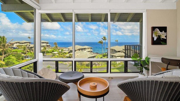 A balcony with wicker furniture, a wooden table, and a view of the ocean and buildings. - Kapalua Bay Villa 31G1 By Parrish - Great ocean view location with AC (Kapalua)