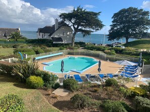A swimming pool with blue and white lounge chairs, surrounded by a white fence, with a view of the sea and houses in the background.