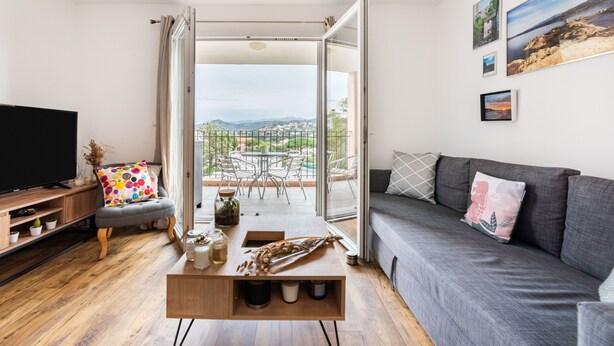 A modern living room with a flat-screen TV, a grey sofa, a wooden coffee table, and a balcony with a view of greenery and buildings. - Occi - proche plage - One Bedroom Apartment, Sleeps 4 (L'Île-Rousse)