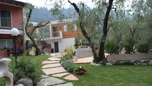 Un sentier de jardin mène à une maison moderne dotée d’un balcon, le tout étant entouré de verdure et de plantes en fleurs.