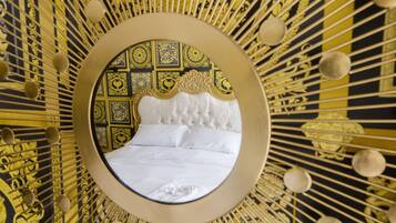 Un espejo circular refleja una cama con cabecera capitonada, rodeada por un muro con diseños dorados y negros.