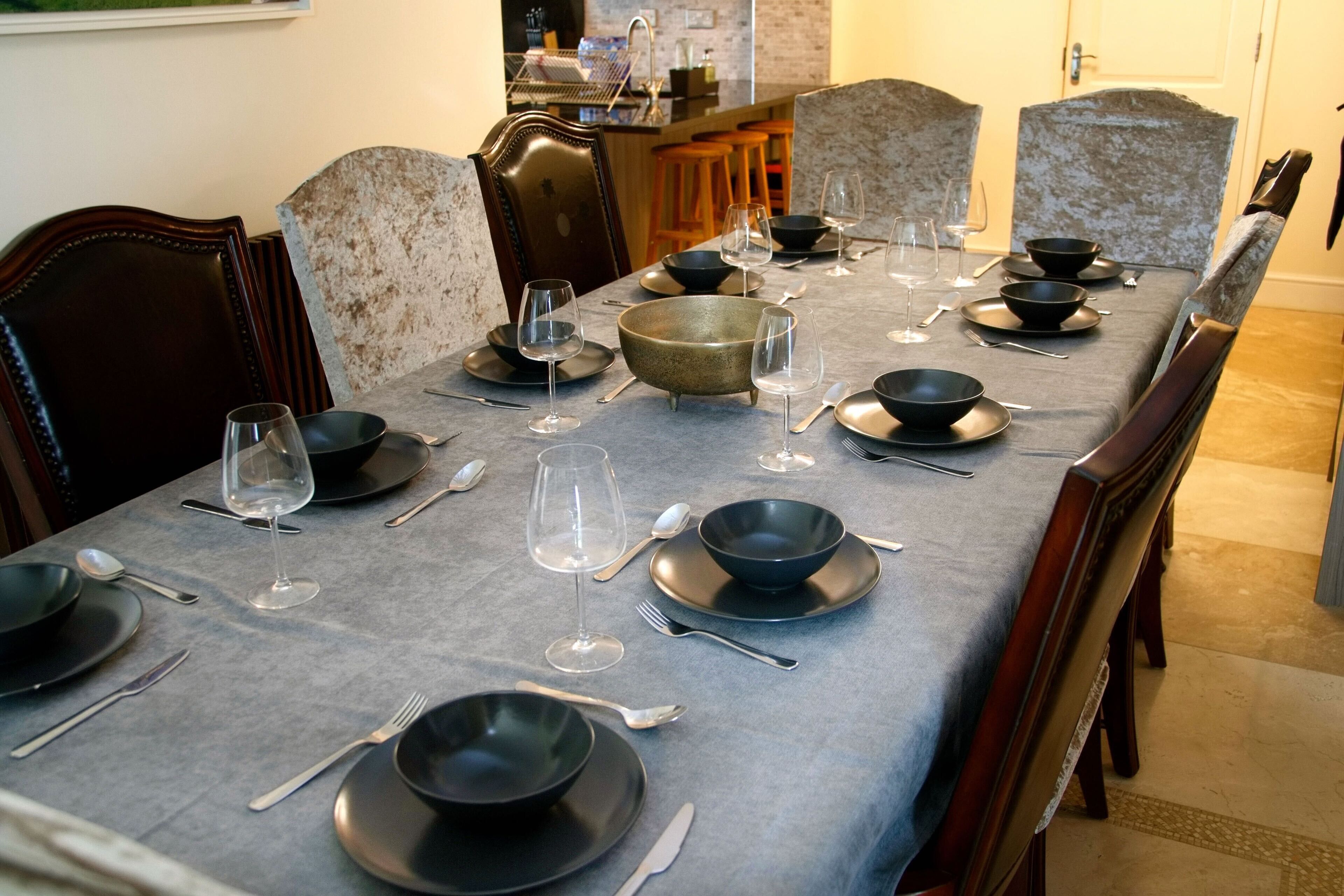 A dining table set with black plates, bowls, and wine glasses, surrounded by upholstered chairs.