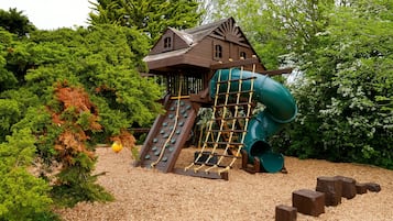 Ein hölzernes Spielhäuschen mit Rutsche und Kletternetz, umgeben von Bäumen und Rindenmulch.