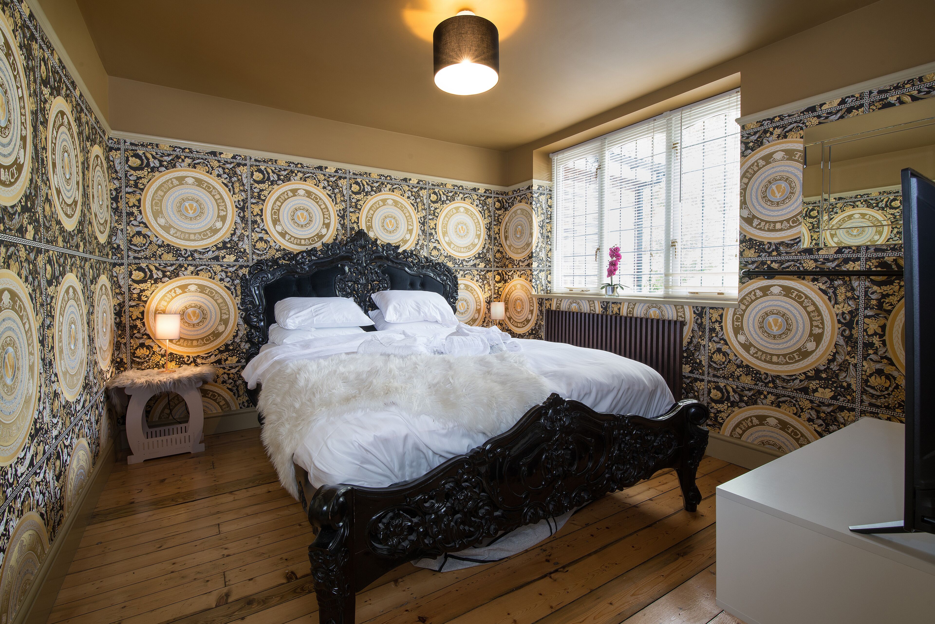 Une chambre avec un grand lit, une petite table de chevet, une fenêtre avec des stores et un papier peint à motifs.