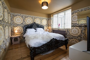 A bedroom with a large bed, a small bedside table, a window with blinds, and patterned wallpaper.