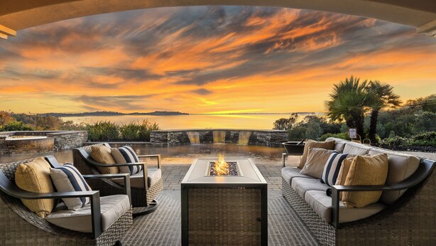 Un área de estar al aire libre con una chimenea, con vistas a un cuerpo de agua y al atardecer.