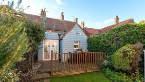 A house with a wooden deck, a front door, and a small garden area with bushes and a pathway leading to the door. - Augusta - cosy cottage near Sheringham town centre (Sheringham)