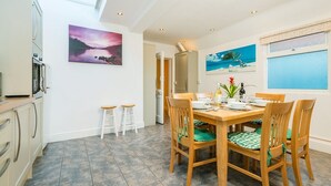 A dining area with a wooden table and chairs, a kitchen area with cabinets and a stove, and two paintings on the walls. - Augusta - cosy cottage near Sheringham town centre (Sheringham)