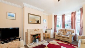 A living room with a fireplace, a sofa, a television, and a large window with curtains. - Augusta - cosy cottage near Sheringham town centre (Sheringham)