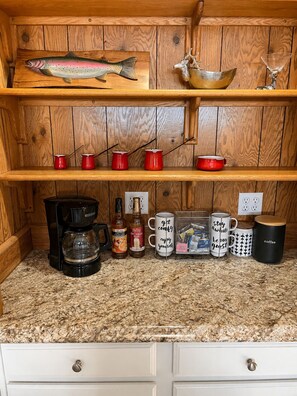 Una cocina con repisas de madera, un adorno de pez, tazas rojas, una cafetera y varias tazas con mensajes.