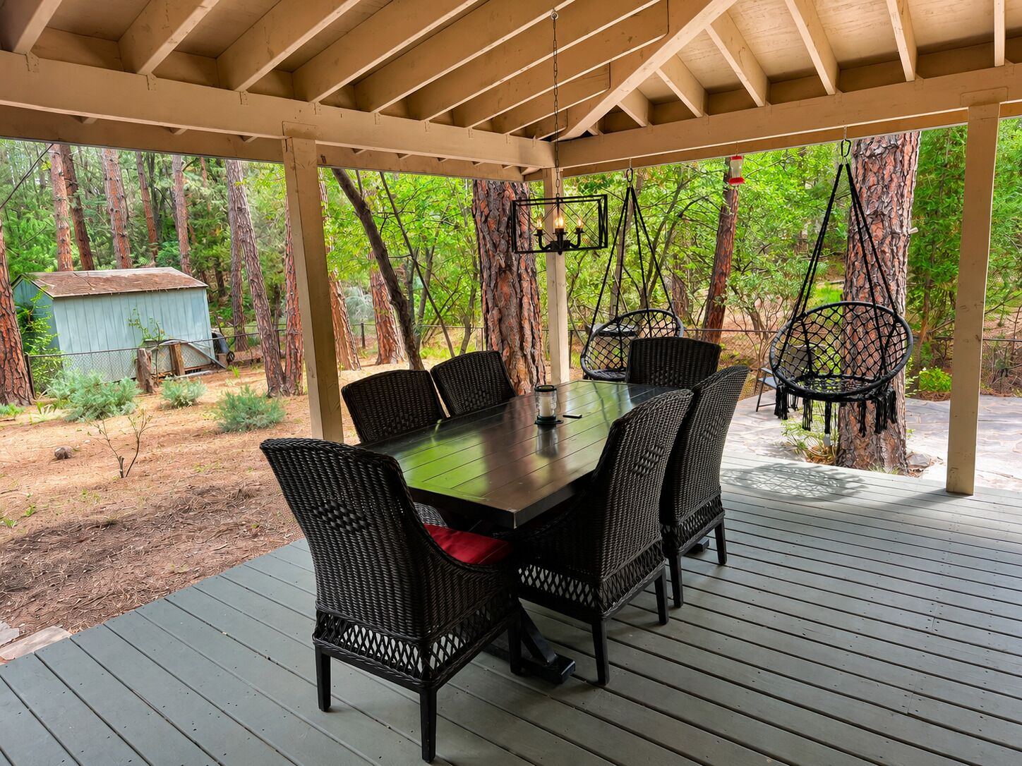 A covered outdoor dining area with a table and chairs, surrounded by trees and a small shed.